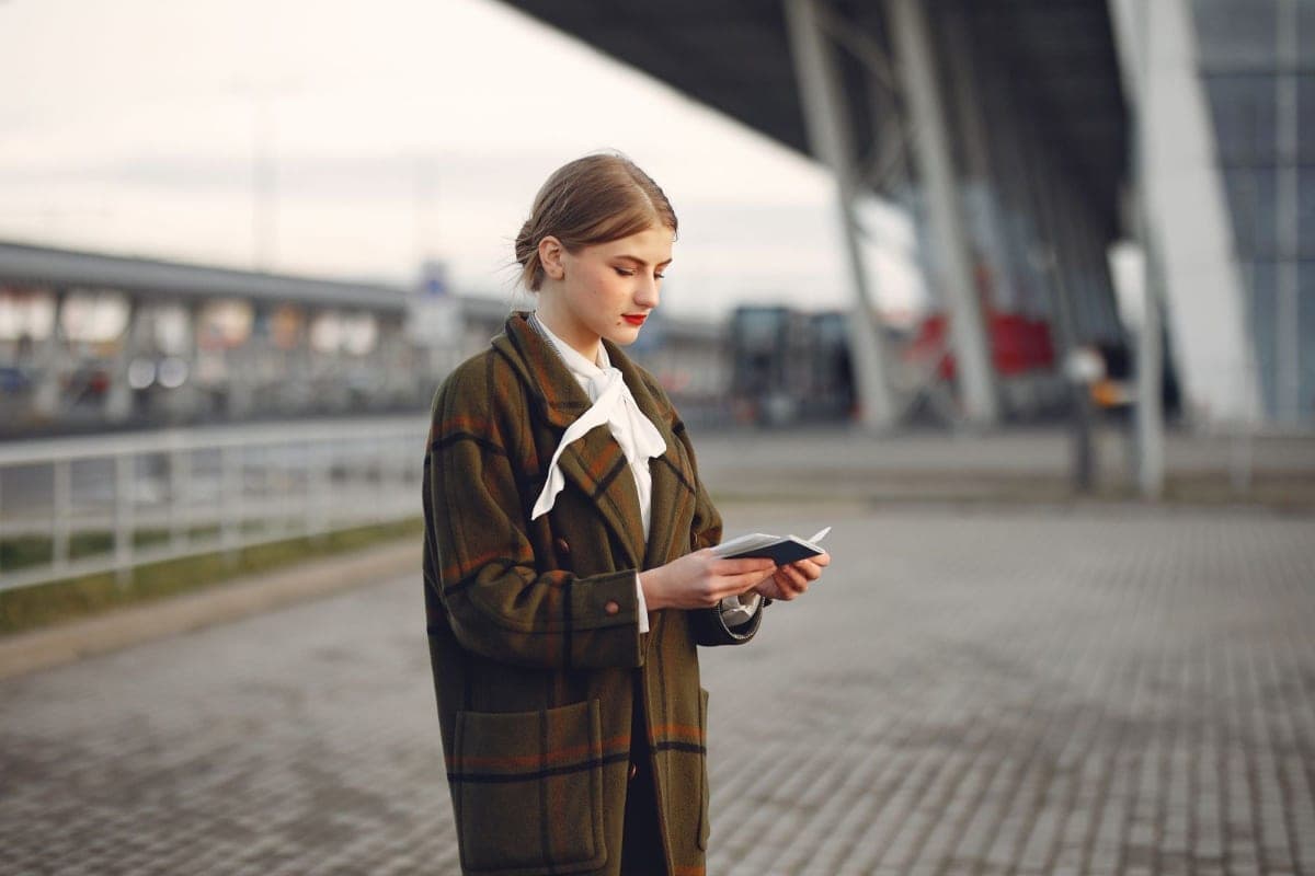 Traveller in a plaid coat checking her passport and boarding pass outside an airport terminal