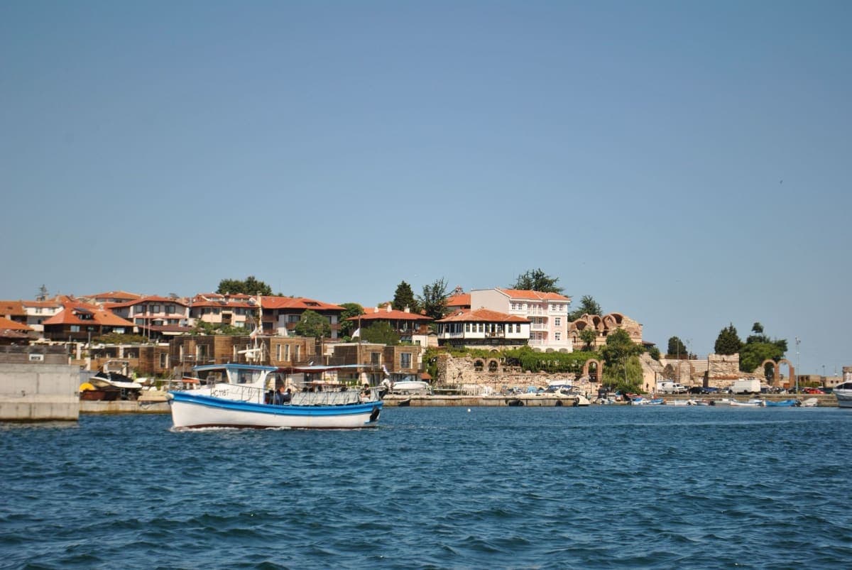 Nessebar's historic waterfront seen from the water, with red-roofed houses, old stone ruins and a white tour boat on the Black Sea